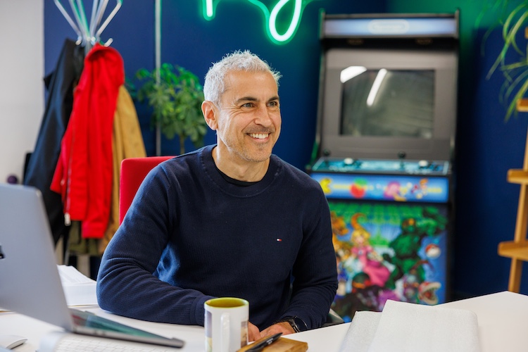 Sponge founder Alex Tosh sits at his desk with his laptop in front of the glowing green Sponge sign and branded arcade machine